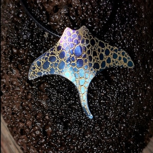 Puede incluir: Un collar con colgante artesanal con un diseño estilizado de manta raya. El colgante presenta un degradado de azules y morados, acentuado con detalles dorados. La forma de la raya se suspende de un cordón negro, creando un contraste llamativo.