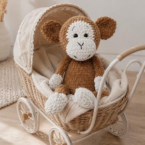 Pode incluir: Um peluche de macaco em croché com fio texturizado castanho e branco, com orelhas grandes e redondas, uma face branca e olhos e boca bordados a preto, sentado dentro de um pequeno carrinho de bebé de vime com rodas brancas e capota.