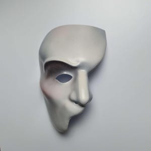Op de afbeelding: Een half-gezichtsmasker met een gladde, lichtgrijze afwerking, een prominente wenkbrauwboog, een gedefinieerde neus en één oogopening. Het ontwerp bevat subtiele airbrush-schaduwen in donkergrijs rond de oogkas en een zachte roze tint.