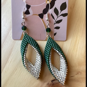 May include: A pair of silver and green beaded earrings with a teardrop shape. The earrings are made with small, square beads and have a dangling green crystal at the top.