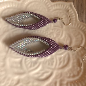 May include: A pair of purple and iridescent beaded earrings with silver hooks. The earrings are shaped like a teardrop with a small hole in the center.