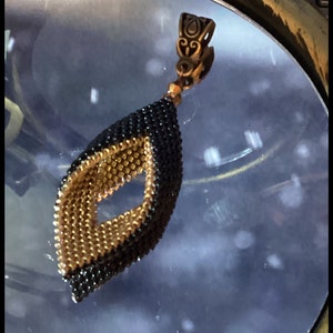 May include: A black and gold beaded pendant with a decorative metal bail. The pendant is shaped like a teardrop with a diamond-shaped opening in the center.