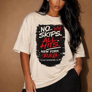 May include: A beige short-sleeve t-shirt featuring a black distressed rectangular graphic with red and white text that reads: NO SKIPS. ALL HITS. NEW YORK R&B EAST RUTHERFORD, NJ. The shirt has a crew neckline and a relaxed, loose-fitting silhouette.