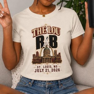 May include: A cream-colored short-sleeve t-shirt featuring a graphic print with the text THE LOU R&B, an illustration of the St. Louis Gateway Arch and a building, and the text ST. LOUIS, MO JULY 21, 2026, paired with blue denim shorts.