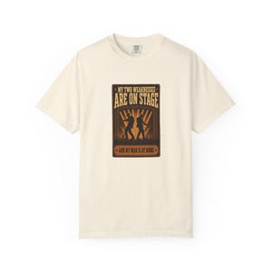 May include: A cream-colored short-sleeve t-shirt featuring a rectangular graphic print. The design displays two silhouettes of performers on a stage with spotlights and the text: MY TWO WEAKNESSES ARE ON STAGE AND MY MAN IS AT HOME.