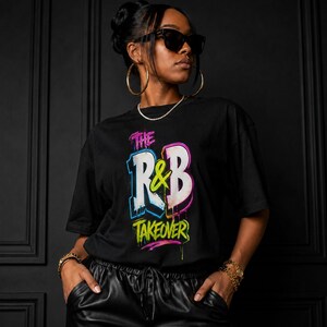 May include: A black short-sleeved t-shirt featuring a graphic print that reads THE R&B TAKEOVER in stylized, multi-colored neon-effect lettering with pink, blue, yellow, and white accents, set against a solid black background fabric.
