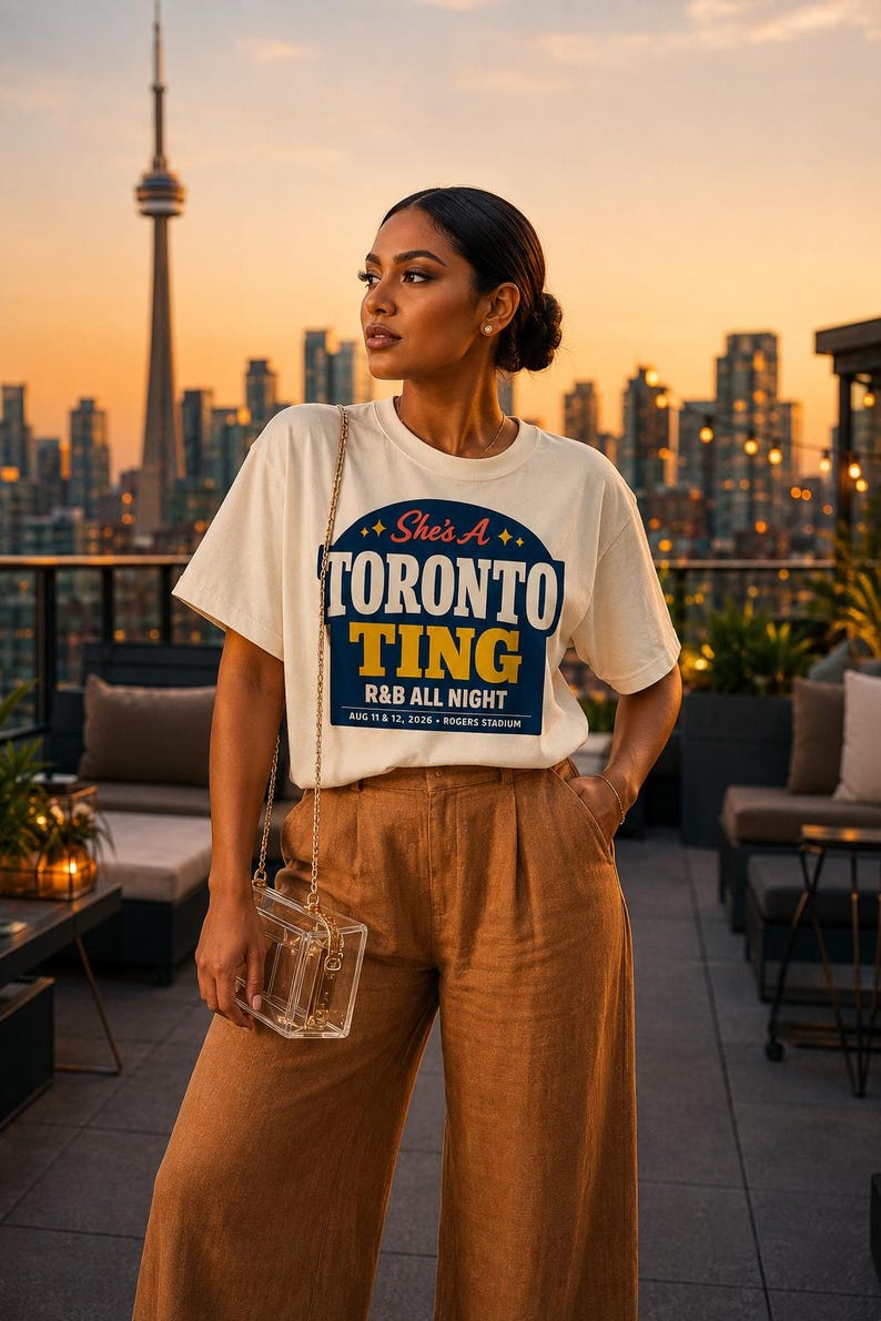 May include: An off-white short-sleeve t-shirt featuring a blue and yellow graphic with the text "She's A TORONTO TING R&B ALL NIGHT AUG 11 & 12, 2026 ROGERS STADIUM" paired with tan high-waisted trousers and a small clear box-style shoulder bag.