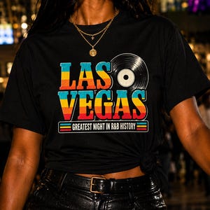 May include: A black short-sleeve t-shirt featuring a graphic print with the text LAS VEGAS in a multi-colored gradient font, a vinyl record icon, and the text GREATEST NIGHT IN R&B HISTORY enclosed in a rectangular border with horizontal stripes.