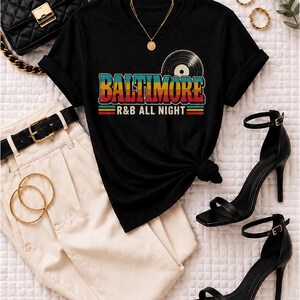 Puede incluir: Una camiseta negra de manga corta con un gr&aacute;fico de un disco de vinilo y el texto BALTIMORE R&B ALL NIGHT en letras de colores retro, combinada con pantalones color crema, un cintur&oacute;n negro, un bolso acolchado negro y sandalias de tac&oacute;n.