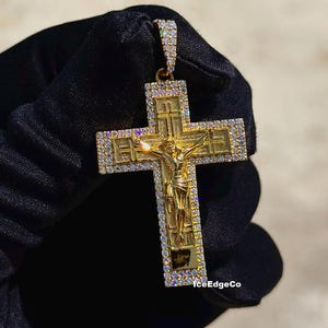 May include: Gold-toned pendant featuring a crucifix design with a detailed depiction of Jesus. The cross is embellished with small, sparkling stones around the edges. The pendant is held against a dark background.