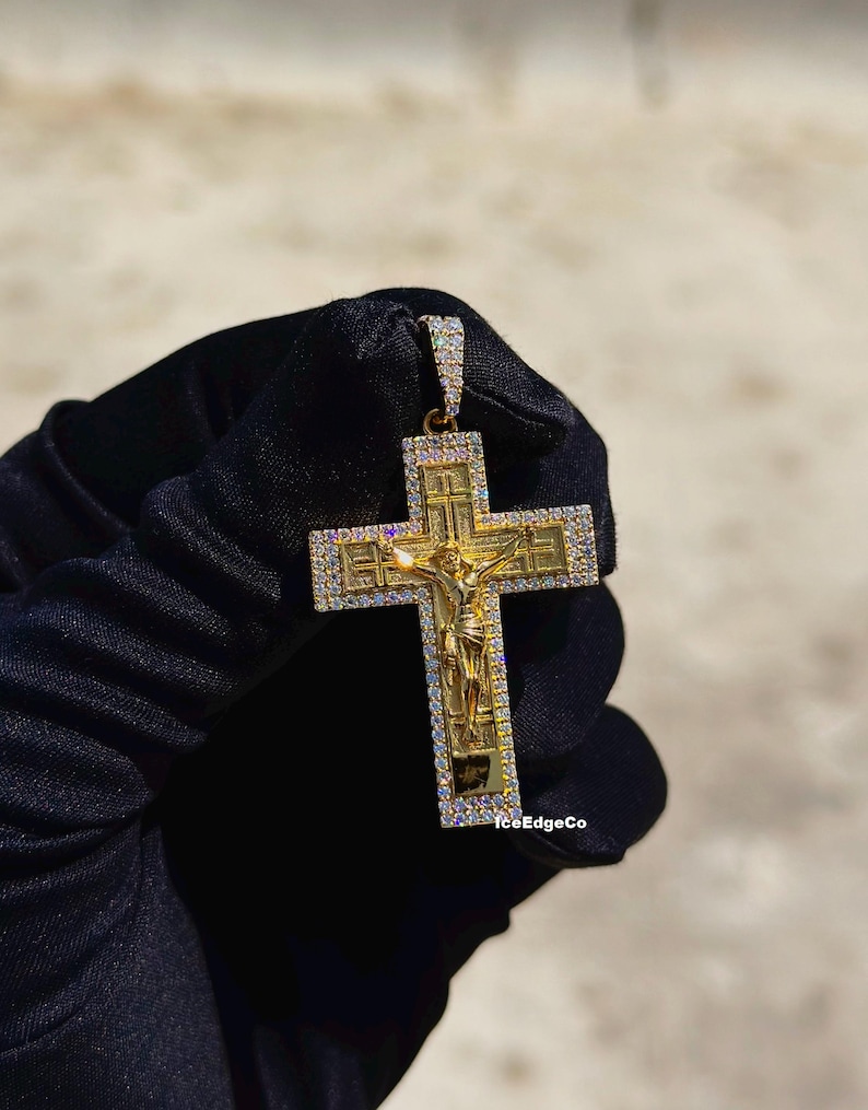 May include: Gold-toned pendant featuring a crucifix design with a detailed depiction of Jesus. The cross is embellished with small, sparkling stones around the edges. The pendant is held against a dark background.