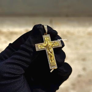 May include: A gold-colored pendant in the shape of a cross, featuring a detailed depiction of Jesus Christ. The cross is adorned with small, sparkling stones. The pendant is held against a dark background.