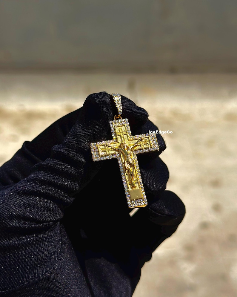 May include: A gold-colored pendant in the shape of a cross, featuring a detailed depiction of Jesus Christ. The cross is adorned with small, sparkling stones. The pendant is held against a dark background.