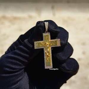 May include: Gold-colored cross pendant with a detailed depiction of Jesus Christ. The cross is encrusted with small, clear gemstones around the edges. The pendant is held against a neutral background. The text "IceedgeCo" is visible on the bottom right.