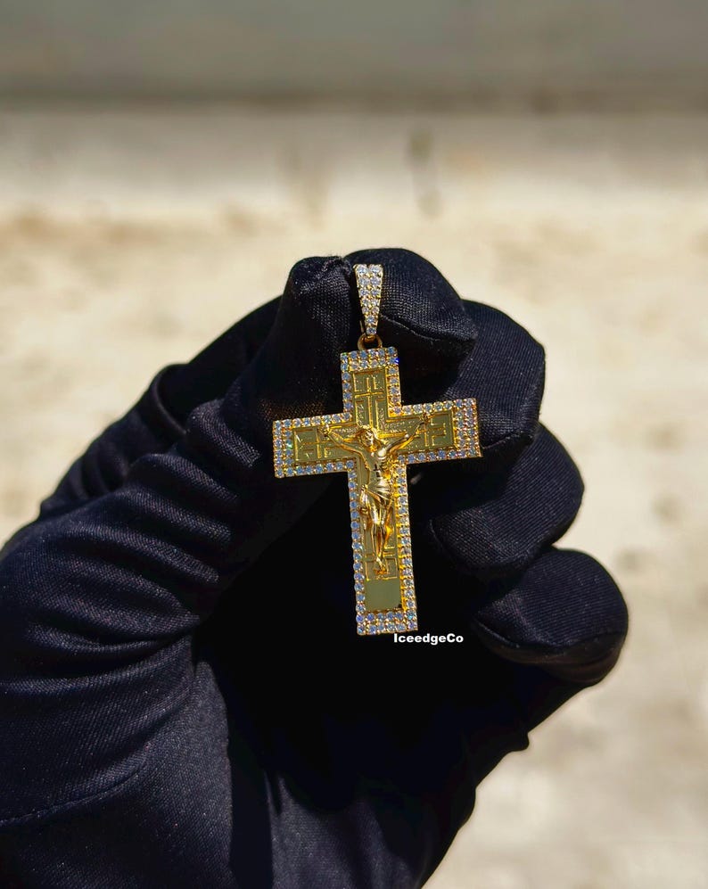 May include: Gold-colored cross pendant with a detailed depiction of Jesus Christ. The cross is encrusted with small, clear gemstones around the edges. The pendant is held against a neutral background. The text "IceedgeCo" is visible on the bottom right.