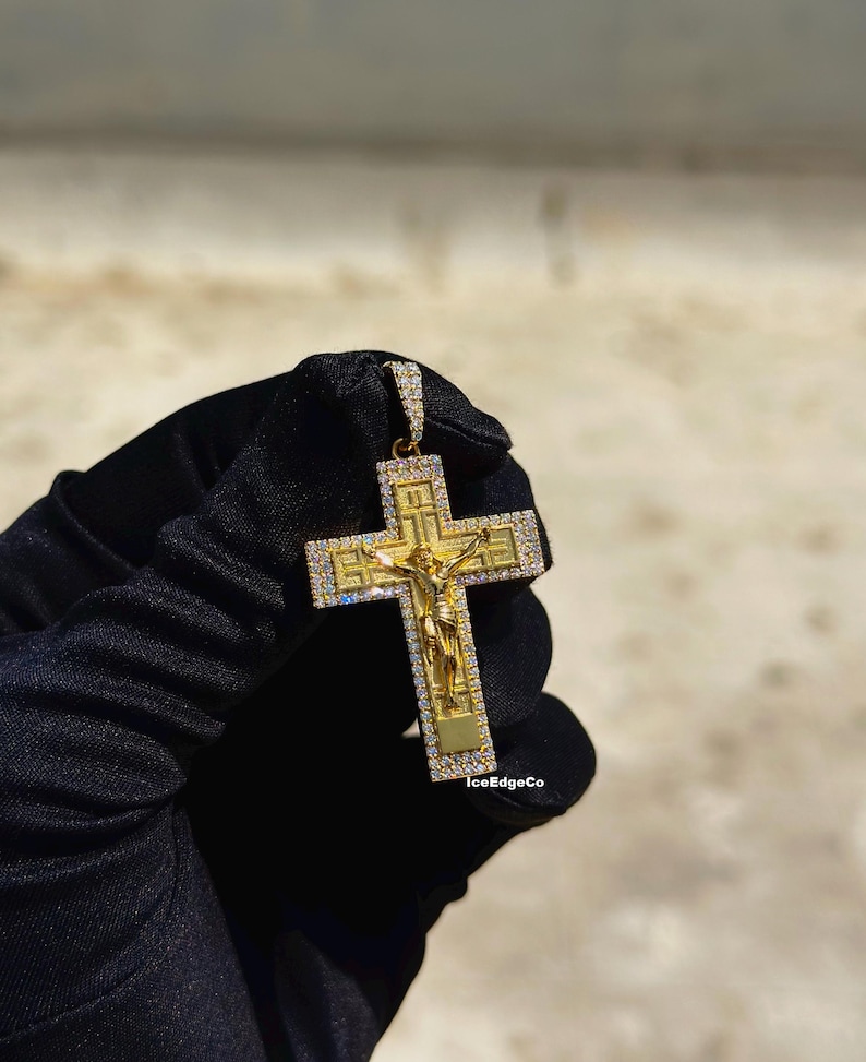 May include: A gold-colored pendant in the shape of a cross, featuring a depiction of Jesus Christ. The cross is adorned with small, clear gemstones and is held by a gloved hand. The text "IceEdgeCo" is visible.