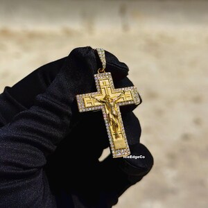 May include: Gold-colored pendant featuring a crucifix design with a detailed depiction of Jesus. The cross is encrusted with small, sparkling stones. The pendant is held against a neutral background.