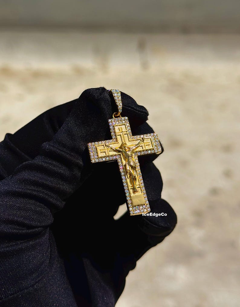 May include: Gold-colored pendant featuring a crucifix design with a detailed depiction of Jesus. The cross is encrusted with small, sparkling stones. The pendant is held against a neutral background.