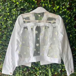 Könnte beinhalten: Weiße Jeansjacke mit einem durchsichtigen Überzug mit gestickten Gänseblümchen. Die Jacke hat ein klassisches Design mit Knopfleiste und langen Ärmeln.