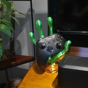 Peut inclure: Une main verte imprimée en 3D tenant une manette Xbox noire. La main repose sur une surface en bois avec un haut-parleur noir en arrière-plan.