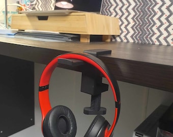 May include: A black and red headphone holder with a clamp that attaches to a desk or table. The holder is designed to hold headphones securely and keep them organized.