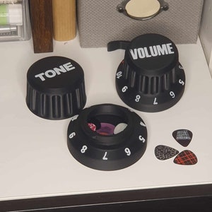 Peut inclure: Trois boutons en plastique noir avec des lettres blanches. Les boutons sont étiquetés "TONE", "VOLUME" et "6 7 8 9 10". Les boutons sont sur une surface blanche avec des médiators de guitare éparpillés autour d'eux.