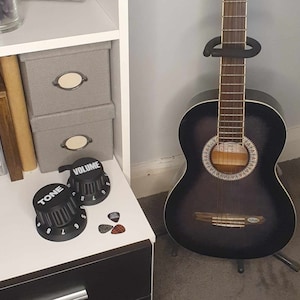 May include: A black acoustic guitar with a wood neck and silver tuning pegs. The guitar is on a stand and has a volume and tone knob.