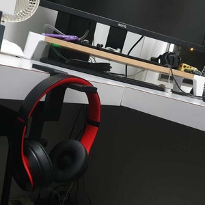 Peut inclure: Casque audio noir avec des accents rouges accroché à un crochet de bureau. Le casque est sur un bureau blanc avec un écran d'ordinateur noir en arrière-plan.