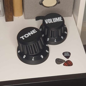 May include: Two black knobs with white lettering, one labeled "TONE" and the other "VOLUME". Both knobs have numbers 5, 6, 7, 8, 9, and 10 printed on them. The knobs are on a white surface.