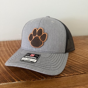 May include: Gray trucker hat with a brown paw print patch. The hat has a black mesh back and a curved brim. The Richardson brand label is visible on the front.