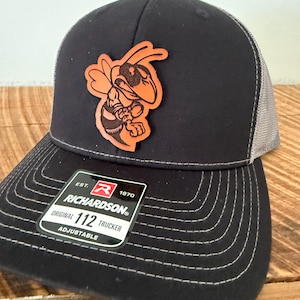 May include: Black and gray Richardson trucker hat with a leather patch of a cartoon hornet. The hat has a curved brim and an adjustable strap. The patch is orange and black. The tag reads "Richardson Original 112 Trucker Adjustable."