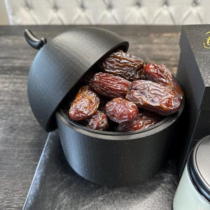May include: A black ceramic bowl with a lid shaped like a dome, filled with dried dates. The bowl is sitting on a dark wooden surface.