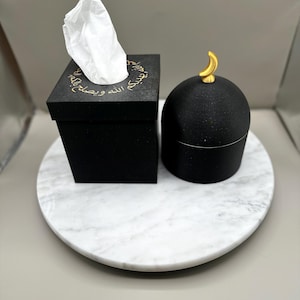 May include: A black tissue box with gold Arabic script and a white tissue sticking out, next to a black, round container with a gold crescent moon on top. Both items sit on a white marble tray.