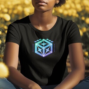 May include: A person wearing a black t-shirt with a colorful geometric design on the front. The design is a cube with a gradient of blue, green, and purple. The person is sitting in a field of yellow flowers.