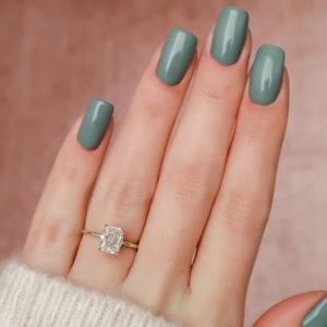 May include: A hand wearing a gold ring with a large, rectangular diamond. The nails are painted a solid, muted green color. The background is a soft, muted pink.