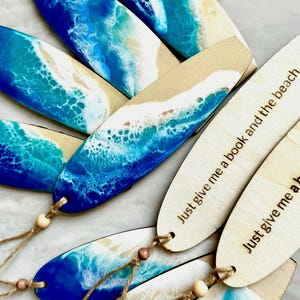 May include: A set of wooden surfboard-shaped bookmarks with blue and white resin art depicting ocean waves. Some bookmarks have the text "Just give me a book and the beach." engraved on them.