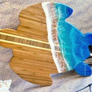 May include: A wooden cutting board shaped like a sea turtle with a blue and white resin ocean wave design. The board has a light brown wood grain and a light brown leather strap.