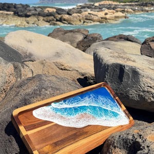 Pu&ograve; includere: Un vassoio da portata in legno con un'intarsio in resina blu e bianca che rappresenta le onde dell'oceano. Il vassoio &egrave; appoggiato su una spiaggia rocciosa.