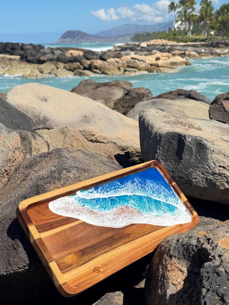 Pu&ograve; includere: Un vassoio da portata in legno con un'intarsio in resina blu e bianca che rappresenta le onde dell'oceano. Il vassoio &egrave; appoggiato su una spiaggia rocciosa.