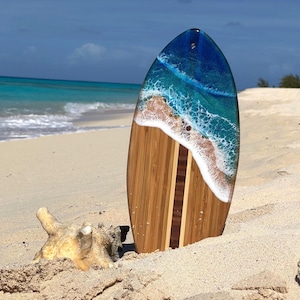 May include: A wooden surfboard with a blue and white epoxy resin ocean wave design. The surfboard is on a sandy beach.