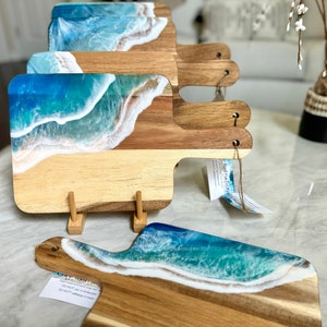 May include: Four wooden cutting boards with a blue and white resin ocean wave design. The boards have a handle and are displayed on a white surface.