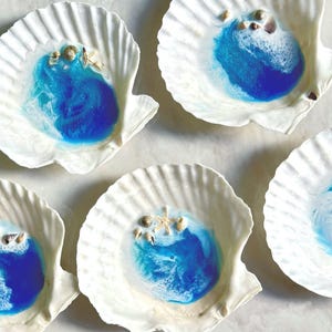 May include: Five white seashells with blue resin inside. The resin has a marbled effect and resembles ocean waves. Each seashell has small white seashells and starfish embedded in the resin.