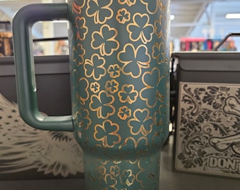 Puede incluir: Una taza de viaje verde con un patrón de trébol dorado. La taza tiene una tapa y un asa.