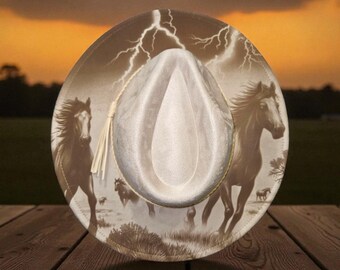 May include: A cowboy hat with a cream-colored crown and a wide brim featuring a sepia-toned graphic of running horses and lightning bolts. The hat is set against a sunset backdrop on a wooden surface.
