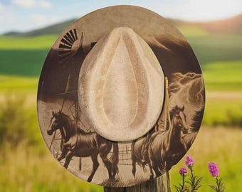 May include: A tan cowboy hat with a detailed illustration of horses, a windmill, and a landscape scene. The hat is resting on a wooden post, with a green field and blue sky in the background.