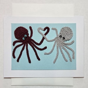 May include: A handmade greeting card featuring two crocheted octopuses. One is burgundy, the other is gray, with their tentacles forming a heart shape. The octopuses have small, round, blue and black eyes, set against a light blue background.