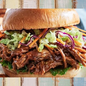 May include: A close-up of a pulled pork sandwich on a white plate. The sandwich has a toasted bun, shredded pork, coleslaw, and a green lettuce base. The sandwich is a hearty meal.