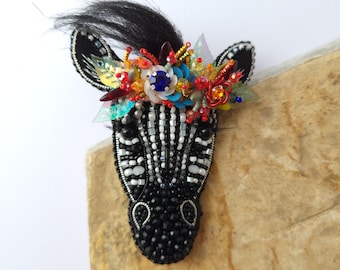 Peut inclure: Broche tête de zèbre faite à la main, ornée de perles noires et blanches. La tête de zèbre est ornée d'une couronne florale colorée avec des perles bleues, rouges et oranges. Une petite touffe de cheveux noirs ajoute une touche fantaisiste à cet accessoire unique.