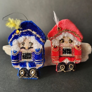 以下が含まれることがあります： ロイヤルブルーと鮮やかな赤のベルベット生地を使用した、刺繍入りのくるみ割り人形のオーナメント2体。各フィギュアには白い髪、金色のメタリック糸のディテール、胸元に黒いビーズのアクセントがあり、帽子には小さな羽が付いています。