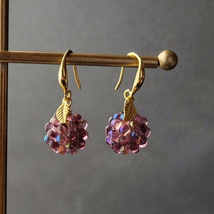 May include: A pair of gold dangle earrings with a cluster of iridescent purple beads shaped like a berry. Each earring has a small gold leaf charm.